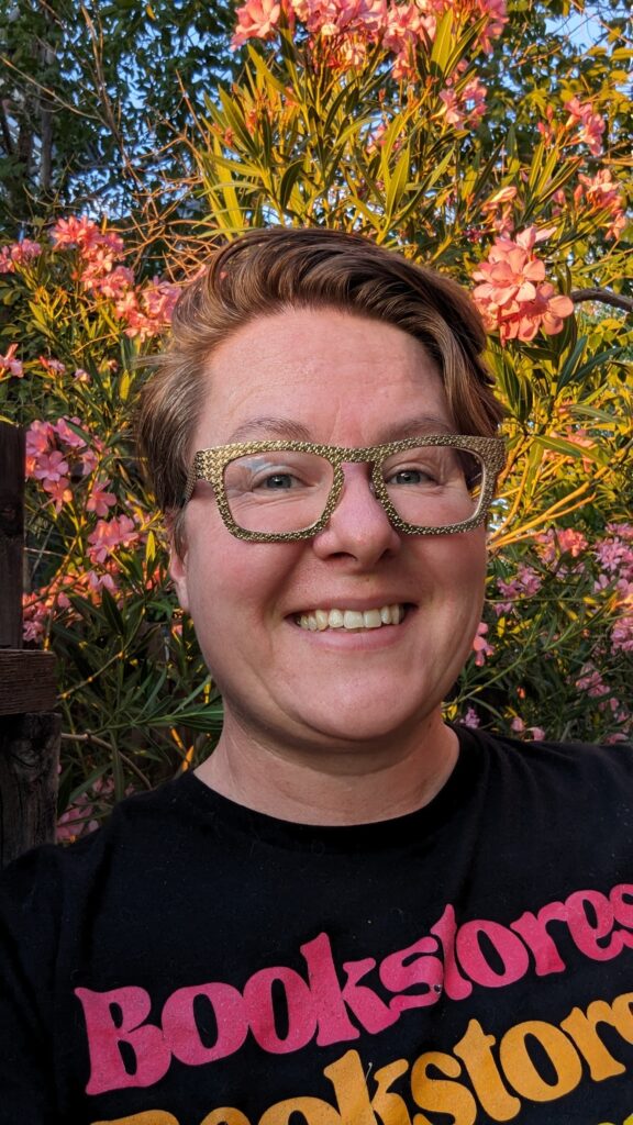 Rath Mercury smiling in front of a flowering oleander bush. Mercury wears gold glasses and a black t-shirt that reads "Bookstores" in rainbow text. They have pale skin with short reddish-brown hair.